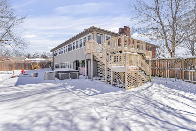 snow covered house with a wooden deck, a fenced backyard, a chimney, stairs, and a hot tub