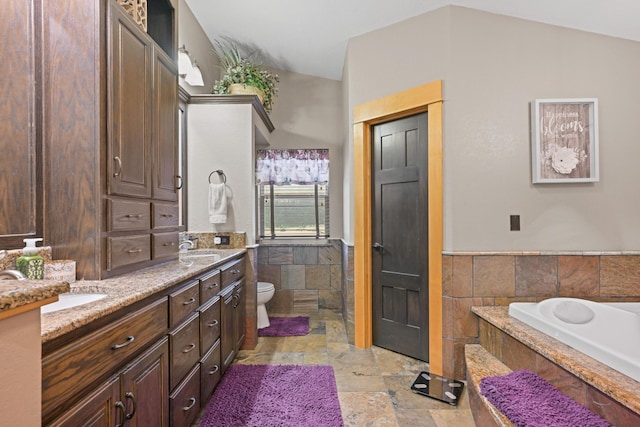 bathroom with a wainscoted wall, tile walls, stone tile floors, double vanity, and toilet