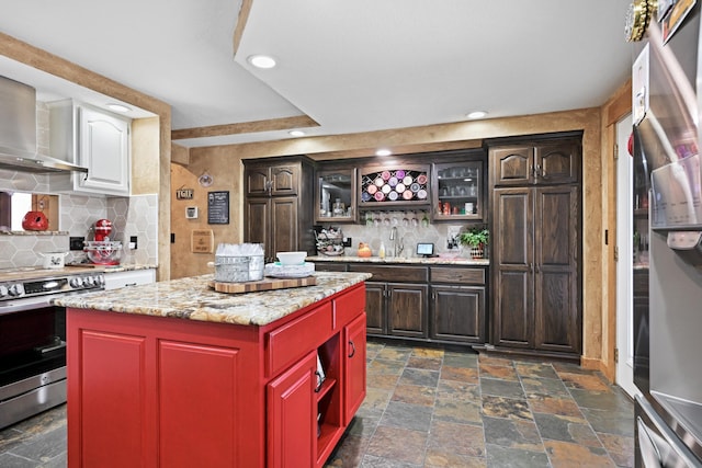 kitchen with glass insert cabinets, a center island, stainless steel appliances, stone tile flooring, and wall chimney range hood