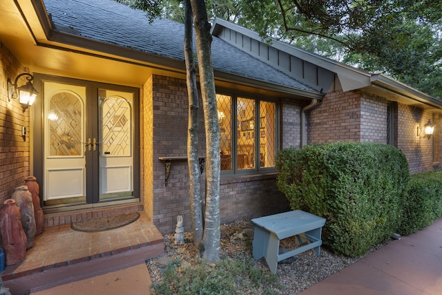 view of exterior entry featuring board and batten siding, a shingled roof, french doors, and brick siding