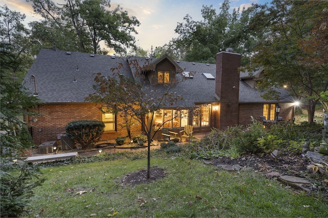 back of property at dusk with a shingled roof, a chimney, a lawn, and brick siding