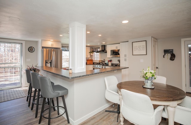 kitchen featuring a kitchen bar, appliances with stainless steel finishes, light wood-style floors, white cabinetry, and wall chimney exhaust hood