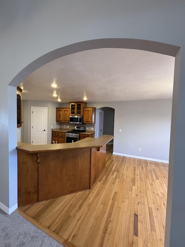 kitchen featuring baseboards, brown cabinetry, arched walkways, a peninsula, and stainless steel appliances