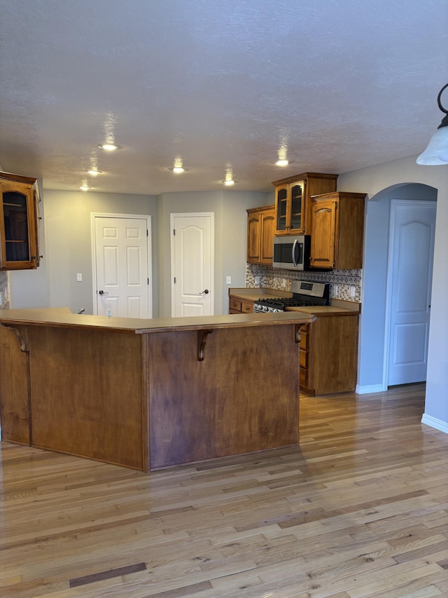 kitchen with tasteful backsplash, arched walkways, glass insert cabinets, stainless steel appliances, and light wood-style floors