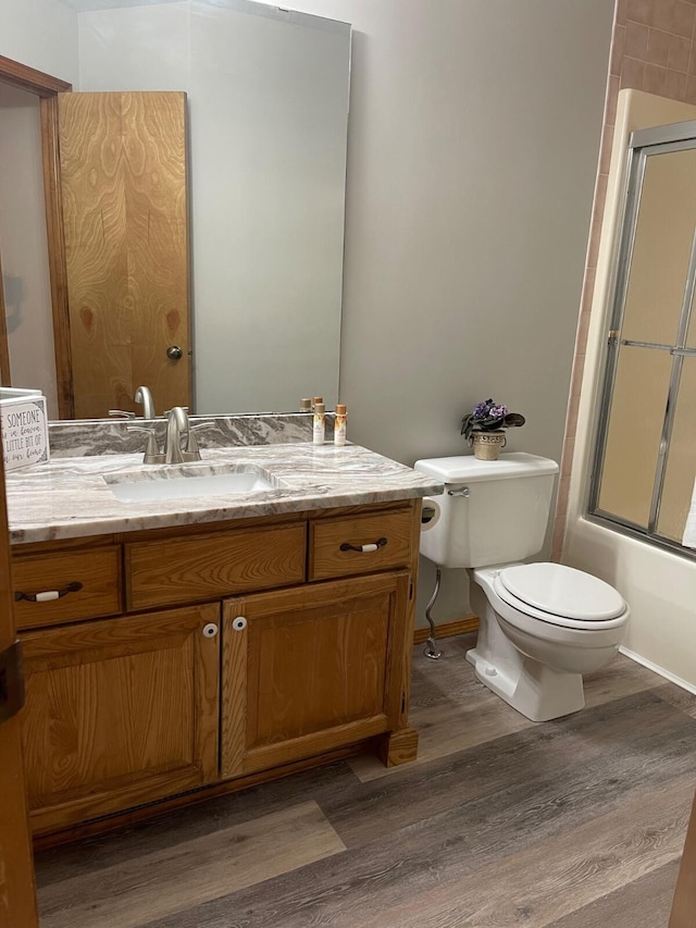 bathroom with toilet, wood finished floors, vanity, and enclosed tub / shower combo