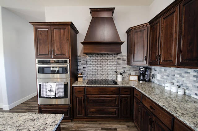 kitchen featuring double oven, black electric stovetop, light stone counters, premium range hood, and decorative backsplash