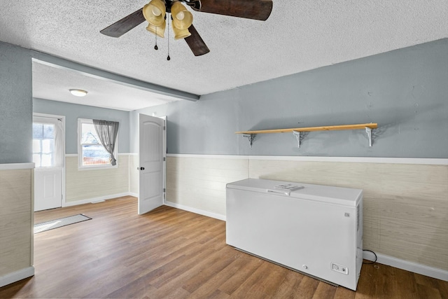 interior space featuring a textured ceiling, a wainscoted wall, wood finished floors, a ceiling fan, and beamed ceiling