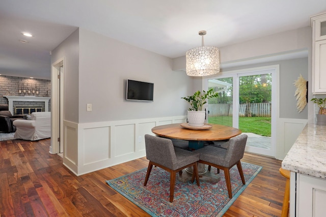 dining space featuring a brick fireplace, a notable chandelier, dark wood finished floors, and a wainscoted wall