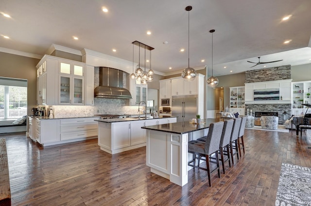 kitchen featuring a large island with sink, a sink, built in appliances, wall chimney exhaust hood, and open floor plan