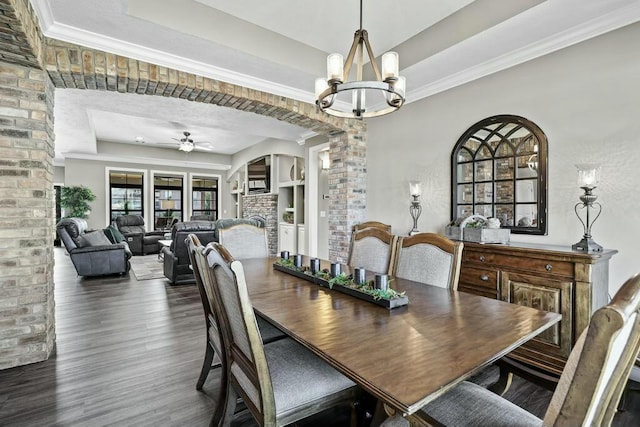 dining area featuring a tray ceiling, dark wood finished floors, ornamental molding, a brick fireplace, and ceiling fan with notable chandelier