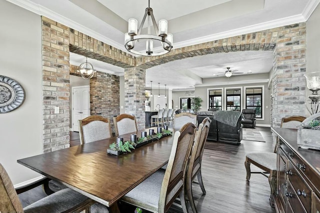 dining room with ceiling fan with notable chandelier, brick wall, dark wood-style flooring, baseboards, and a raised ceiling