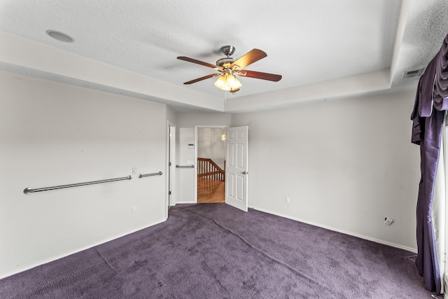 empty room with a tray ceiling, dark carpet, a ceiling fan, a textured ceiling, and baseboards