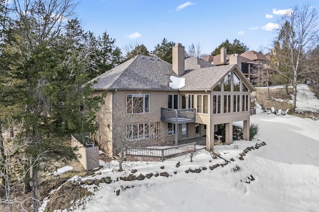 snow covered back of property featuring a shingled roof, a sunroom, a chimney, and stucco siding