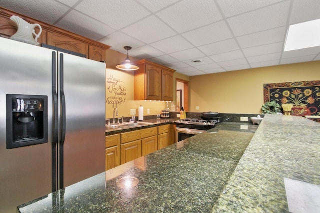 kitchen with a drop ceiling, stainless steel appliances, a sink, brown cabinetry, and dark stone countertops