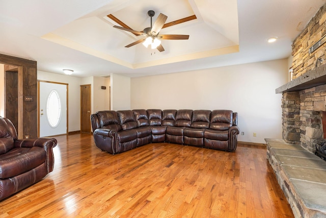 living room featuring ceiling fan, a stone fireplace, a tray ceiling, and light wood-style flooring