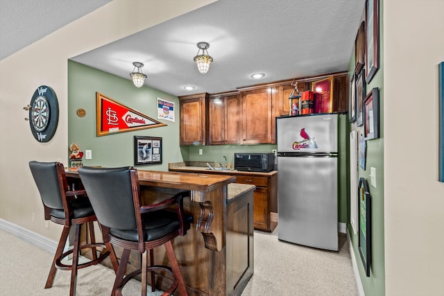 kitchen with brown cabinets, freestanding refrigerator, a textured ceiling, black microwave, and baseboards