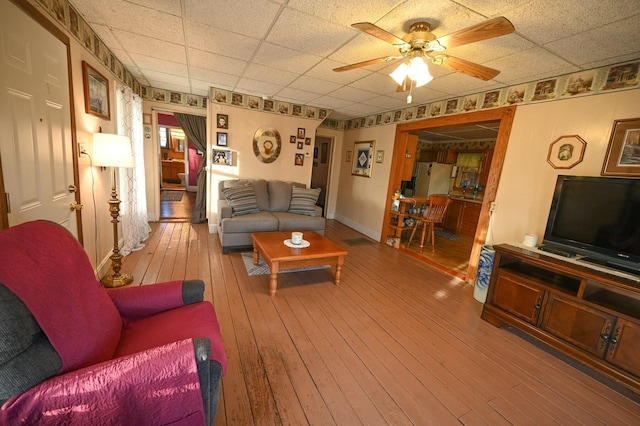 living area with baseboards, a drop ceiling, hardwood / wood-style flooring, and a ceiling fan