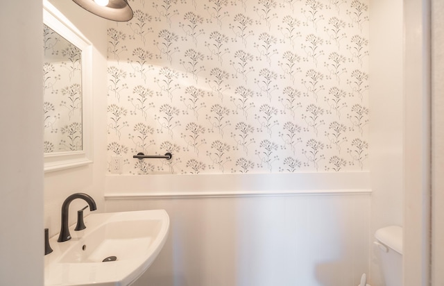 half bath with toilet, wallpapered walls, a sink, and wainscoting