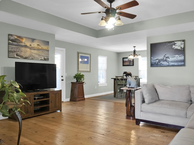 living room featuring light wood finished floors, baseboards, a tray ceiling, and ceiling fan with notable chandelier