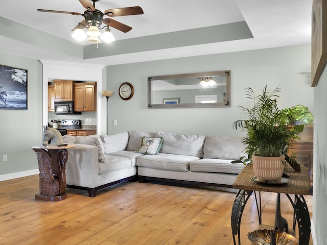 living area featuring light wood finished floors, ceiling fan, a tray ceiling, and baseboards