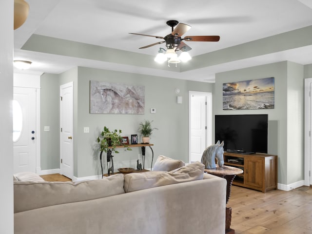 living room featuring ceiling fan, a tray ceiling, light wood finished floors, and baseboards