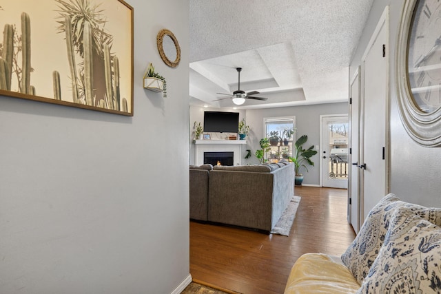 living room featuring a tray ceiling, ceiling fan, a textured ceiling, wood finished floors, and a lit fireplace