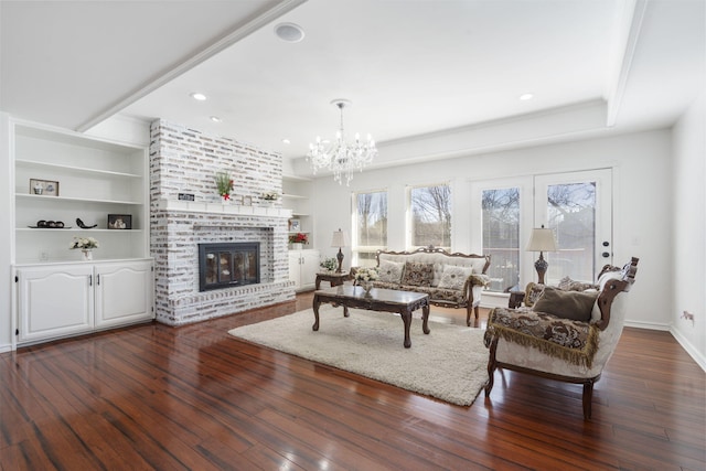 living area with baseboards, built in features, dark wood-type flooring, a brick fireplace, and a chandelier