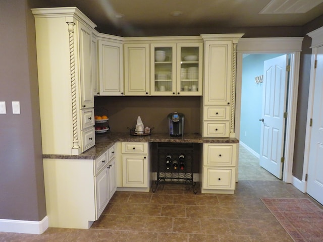 kitchen featuring glass insert cabinets, cream cabinetry, baseboards, and dark stone countertops