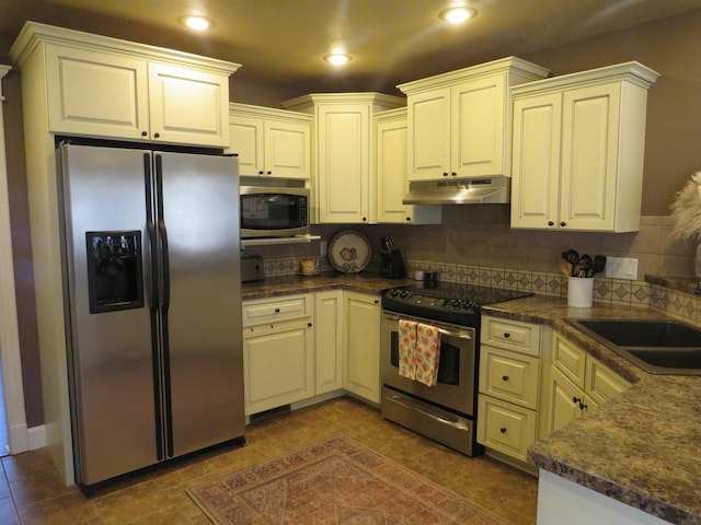 kitchen featuring tasteful backsplash, dark countertops, appliances with stainless steel finishes, under cabinet range hood, and a sink