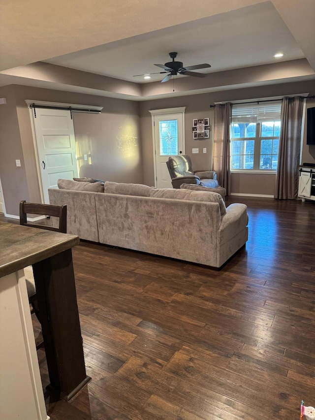 living room with ceiling fan, a barn door, dark wood-style flooring, and a raised ceiling