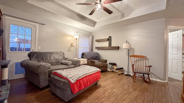 living room with a textured ceiling, ceiling fan, wood finished floors, baseboards, and a tray ceiling