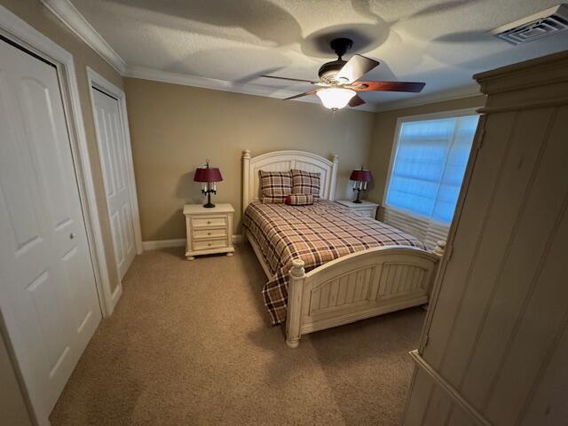 carpeted bedroom with a textured ceiling, ceiling fan, visible vents, two closets, and crown molding