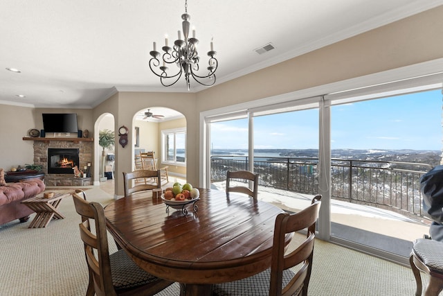 carpeted dining area with arched walkways, crown molding, a fireplace, visible vents, and ceiling fan with notable chandelier