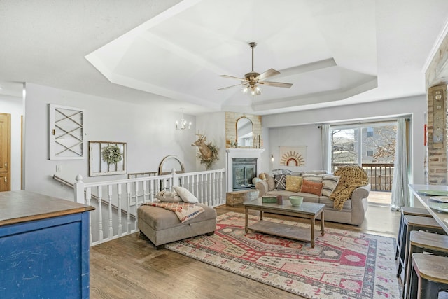 living area with ceiling fan with notable chandelier, a tray ceiling, a fireplace, and wood finished floors