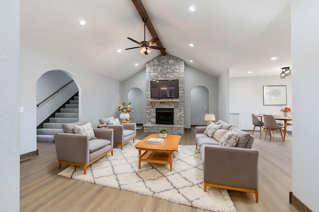 living room featuring a ceiling fan, wood finished floors, stairs, a stone fireplace, and beam ceiling