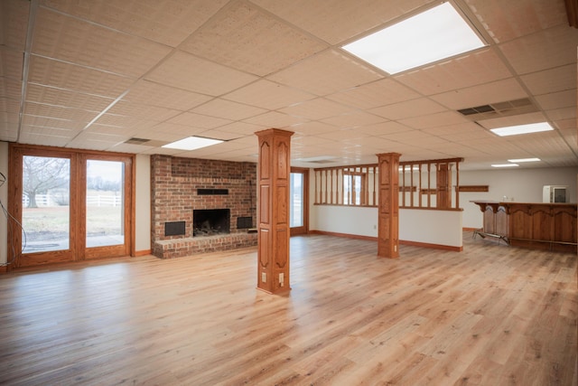 unfurnished living room with decorative columns, light wood finished floors, a brick fireplace, a drop ceiling, and baseboards