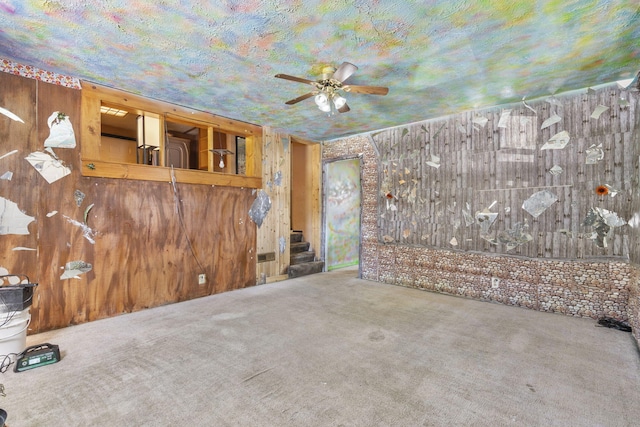 carpeted empty room with stairs, ceiling fan, and wooden walls