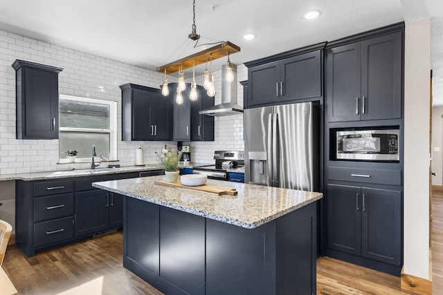 kitchen featuring wood finished floors, light stone countertops, stainless steel appliances, wall chimney range hood, and backsplash