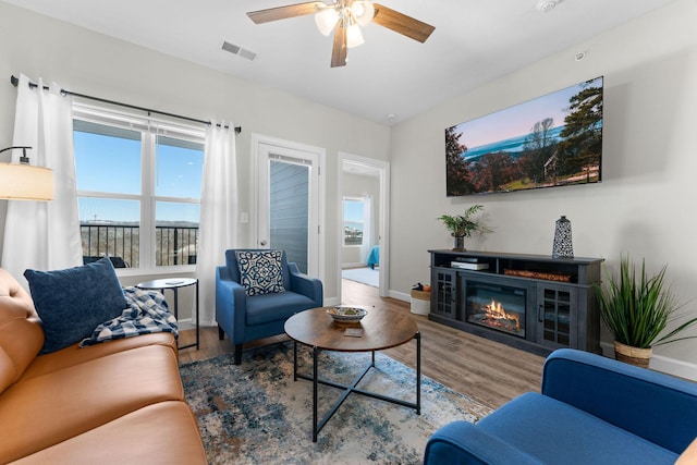 living area with a ceiling fan, visible vents, wood finished floors, and a glass covered fireplace