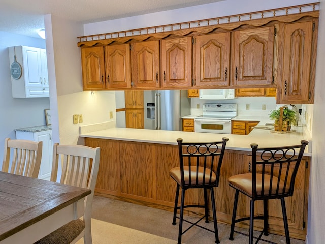 kitchen with light countertops, brown cabinetry, white appliances, a peninsula, and a kitchen breakfast bar