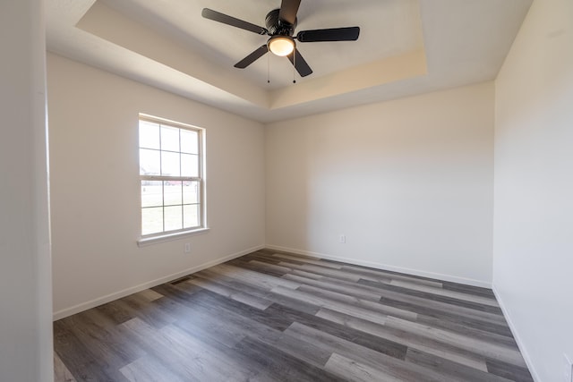 empty room with a tray ceiling, visible vents, dark wood-type flooring, a ceiling fan, and baseboards