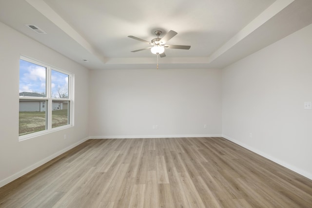 empty room with visible vents, light wood-style flooring, a raised ceiling, and baseboards