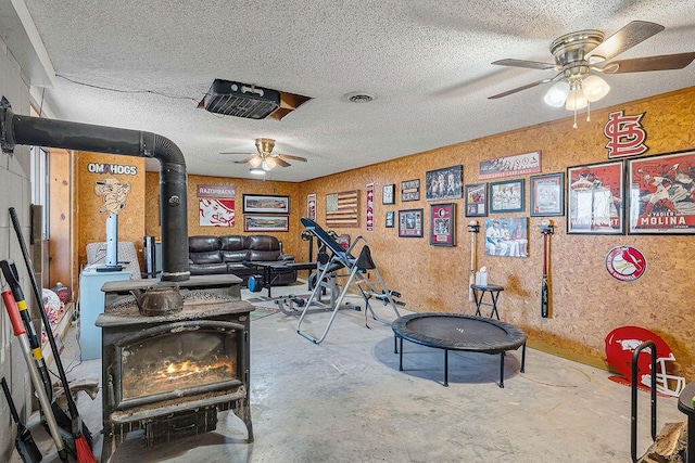 recreation room with unfinished concrete flooring, visible vents, a wood stove, ceiling fan, and a textured ceiling