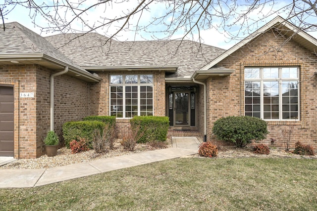 doorway to property featuring a yard, a shingled roof, an attached garage, and brick siding