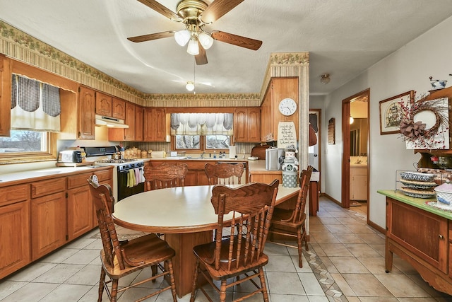 kitchen featuring light tile patterned floors, under cabinet range hood, light countertops, brown cabinetry, and gas range