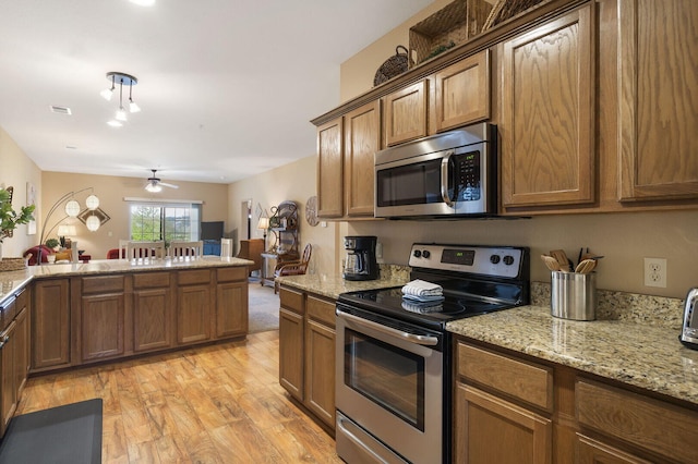 kitchen with light wood finished floors, appliances with stainless steel finishes, brown cabinetry, and light stone countertops