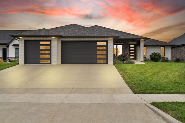 prairie-style home with brick siding, a shingled roof, concrete driveway, a lawn, and an attached garage