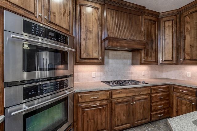 kitchen with decorative backsplash, light stone countertops, stone finish flooring, custom exhaust hood, and stainless steel appliances