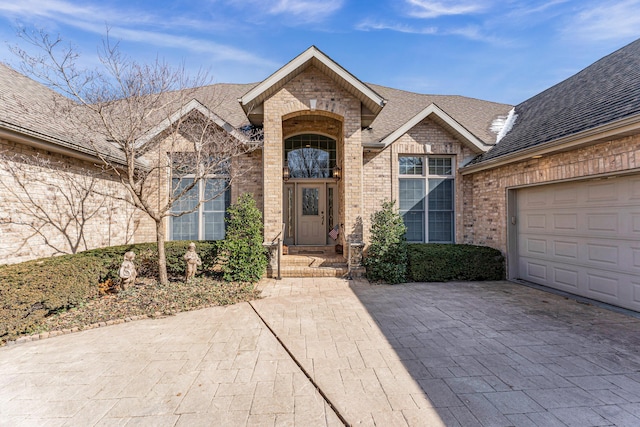 entrance to property featuring a garage, a shingled roof, decorative driveway, and brick siding