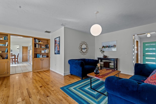 living area with a textured ceiling, built in shelves, visible vents, baseboards, and light wood-style floors